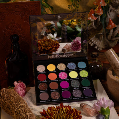 Garden of Spells palette open on a spellbook, 20 named pans under a mirrored lid, among dried flowers, a herb bundle and clay mushrooms.