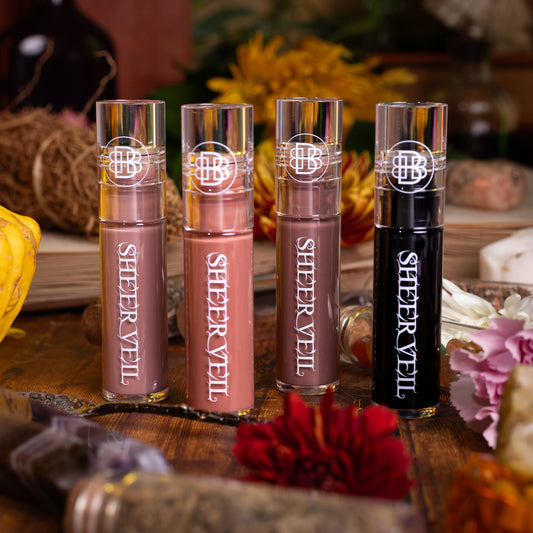 All four Sheer Veil glosses standing together among dried herbs, flowers and jars.
