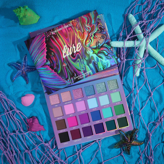 Blend Bunny Cosmetics Lure eyeshadow palette open beside its cover on blue sand, 24 labelled pans in four rows with shells and a fishing net around it