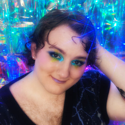 Miles of Multichromatica wearing a Cosmic Cove eye look in yellow, teal, and royal blue on a holographic backdrop