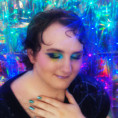 Miles of Multichromatica wearing a Cosmic Cove eye look in green and navy with shimmer, hand at the neck showing teal-blue nail polish