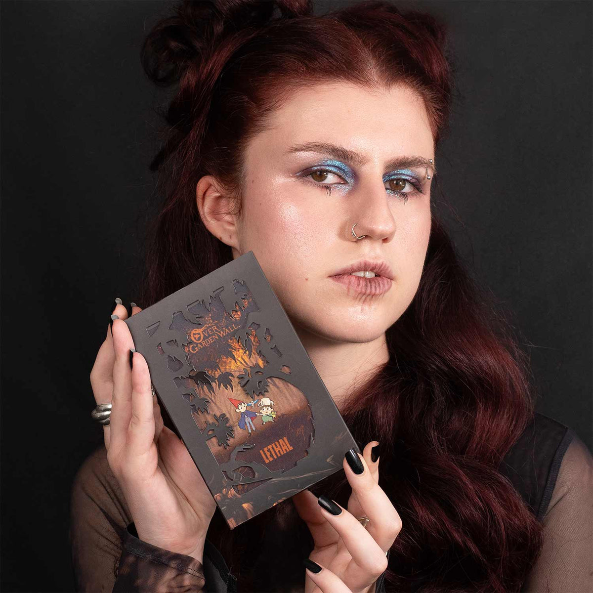 Model holding the closed palette in its laser-cut forest sleeve, wearing a metallic blue eye look