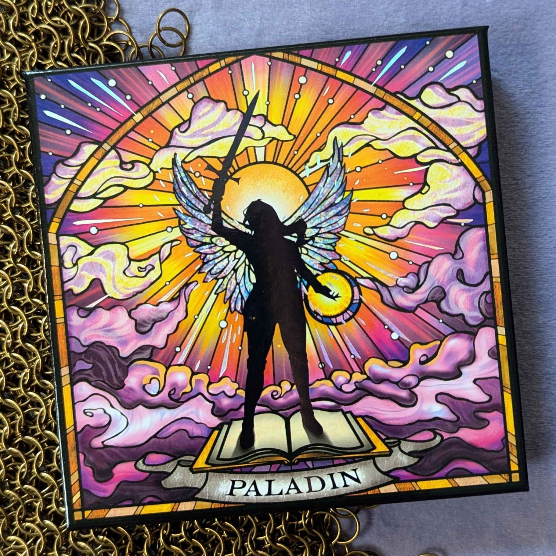 Paladin palette closed on gold chainmail, stained-glass cover art of a winged silhouette holding a sword above an open book, name on a banner below