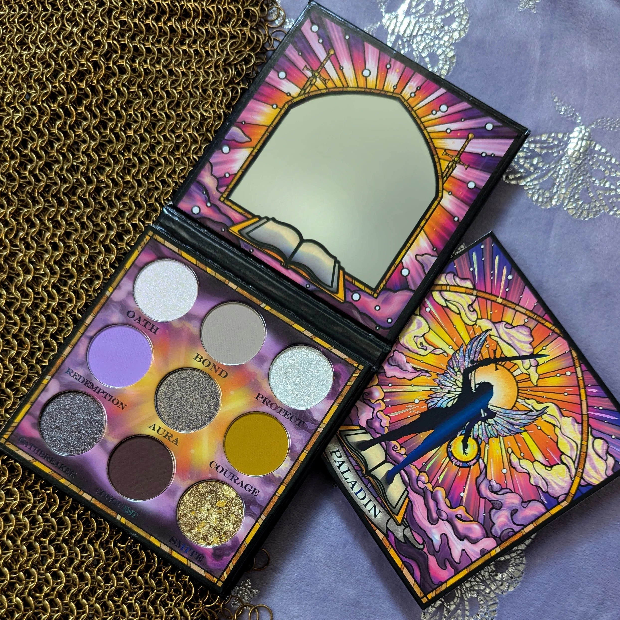 Fantasy Cosmetica Paladin eyeshadow palette open on gold chainmail beside its closed lid, showing nine pans and stained-glass art of a winged sword-bearer
