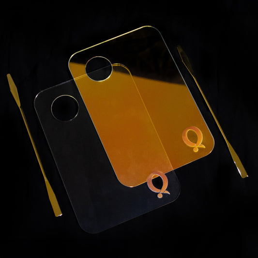 Question Beauty Mixing Palette in clear and holographic gold, both with circle cutouts and gold Q logos, alongside two slim gold spatulas on black.
