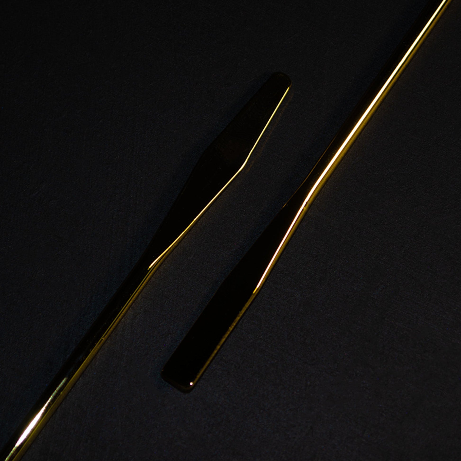 Two slim gold metal mixing spatulas laid diagonally on a dark textured surface.