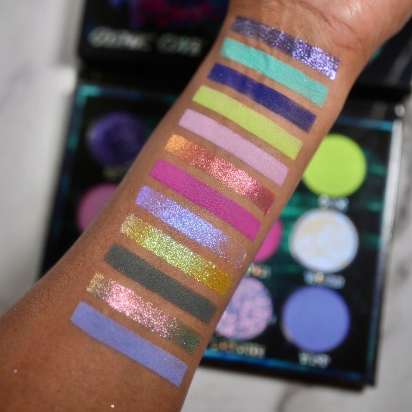 Cosmic Cove swatches on medium skin tone showing all twelve shades in stripes, the open palette visible in the background