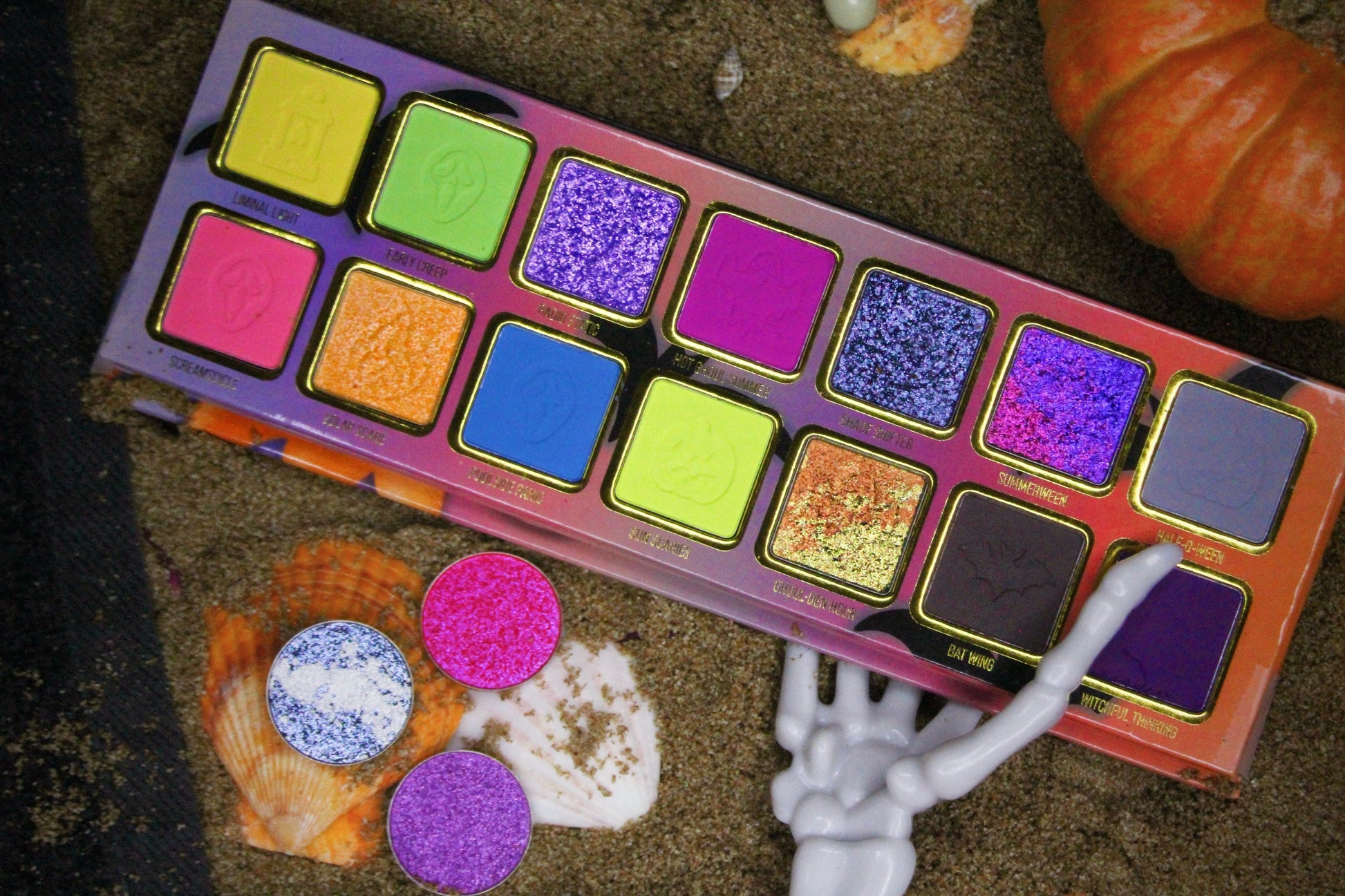Summerween palette in sand with seashells, a small pumpkin, and a skeleton hand, with three loose sparkle pans set in the shells