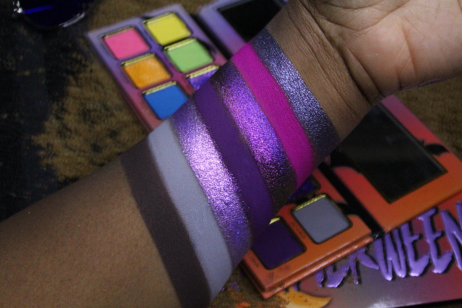 Purple and magenta shifting sparkle shades from Summerween swatched on a deep skin tone, beside soft grey and brown mattes