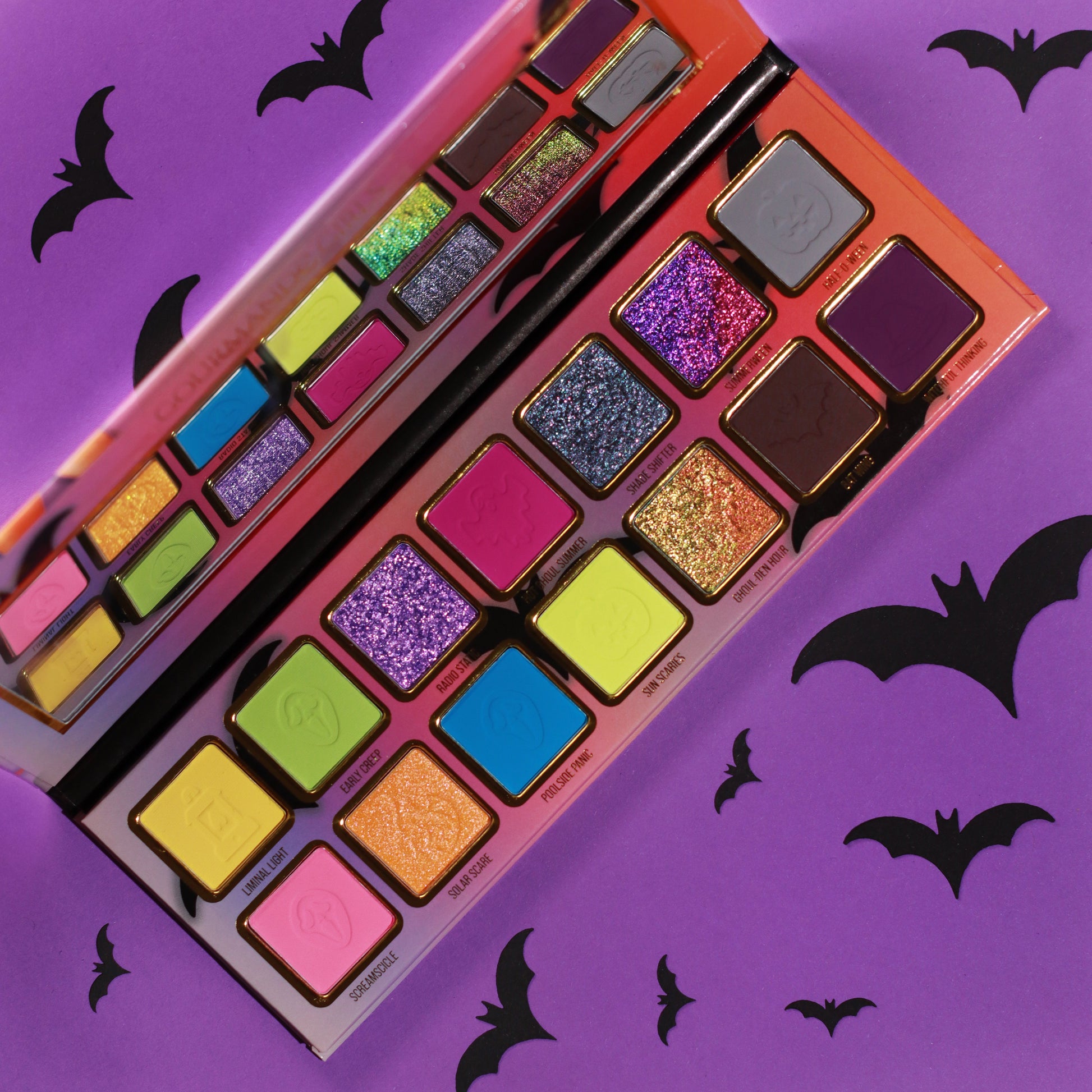 Gourmande Girls Summerween eyeshadow palette open on a purple bat-print background, showing 14 neon and sparkle shades in two rows of gold-framed pans