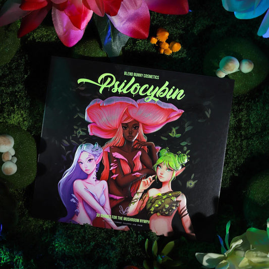 Blend Bunny Cosmetics Psilocybin palette closed, dark box with three mushroom nymph figures in pink, lilac, and green, on a mossy bed with mushrooms and flowers