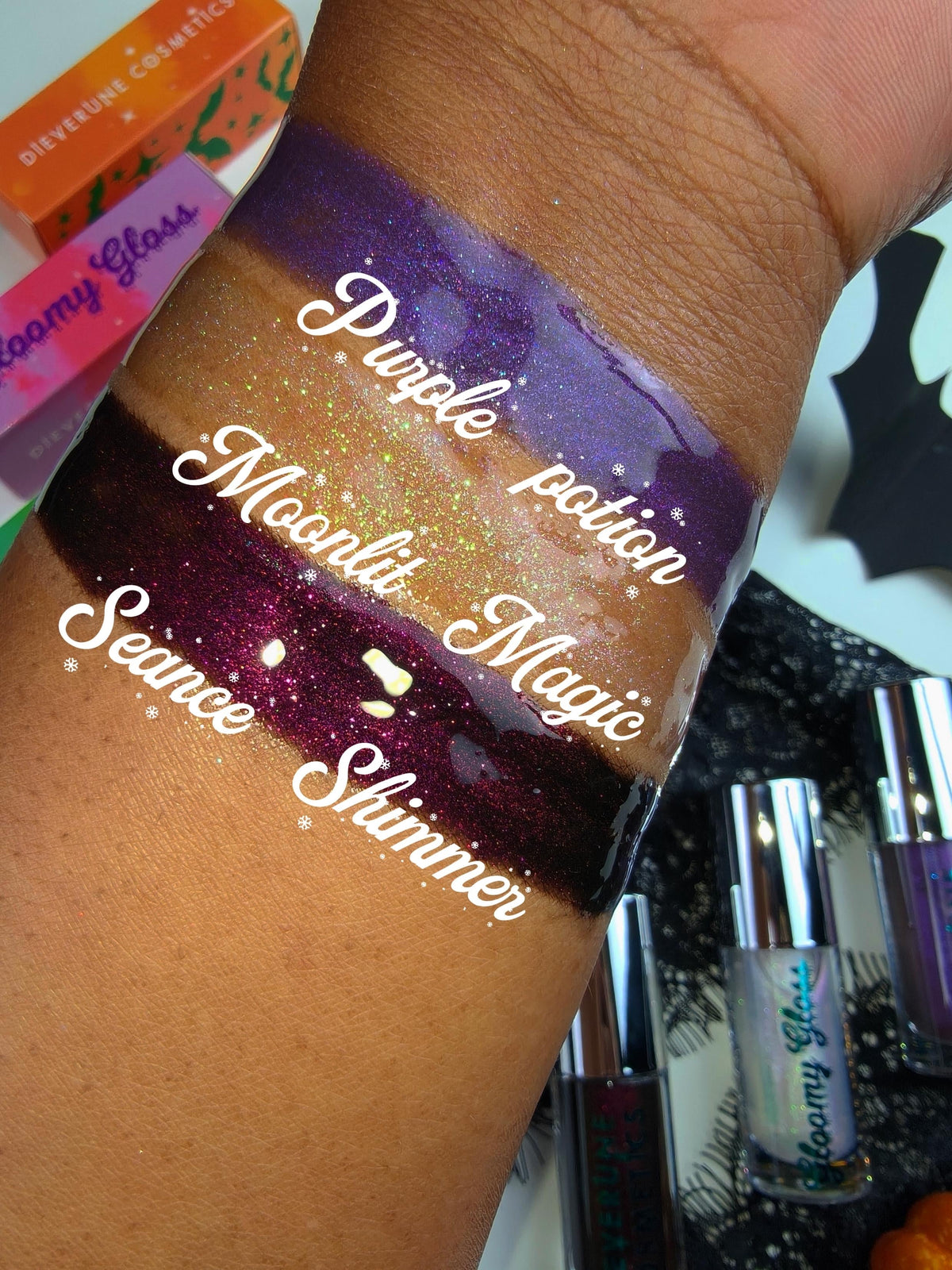 Arm swatches of the three glosses on deep skin, labelled Purple Potion, Moonlit Magic and Seance Shimmer, with the tubes beside them