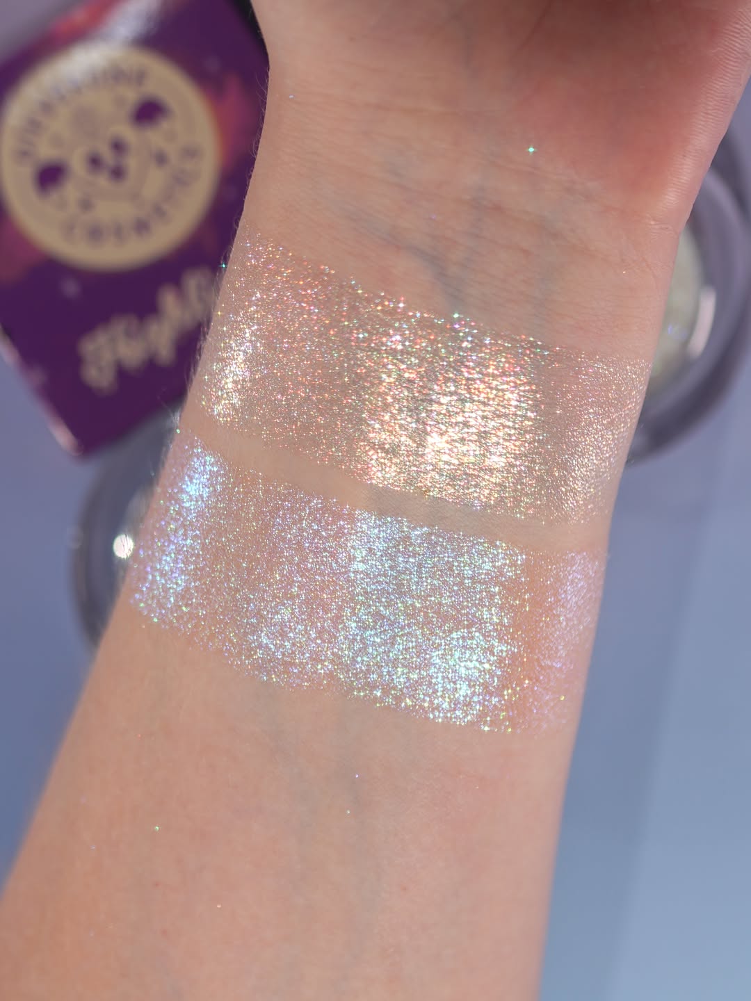 Both highlighters swatched on a fair wrist, a warm sparkle swatch above a blue sparkle swatch, purple Highlighter box behind