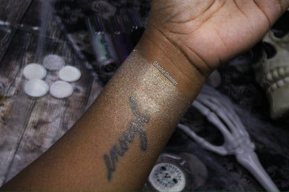 Ghostie Glimmer swatch on deep skin, forearm with a tattoo, skull and skeleton hand props, small brand label