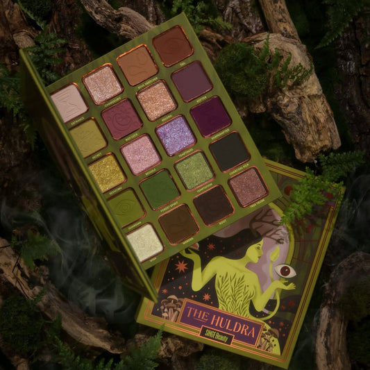 Singe Beauty The Huldra eyeshadow palette open in a mossy forest with smoke, 20 shades across five rows: warm neutrals, greens, plums, and golden sparkles