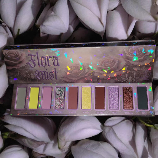 Ladybug Glow Flora Mist eyeshadow palette open among pale lilac petals, holographic rose-print lid above 10 pans of grey, chartreuse, mauve, wine and sparkles
