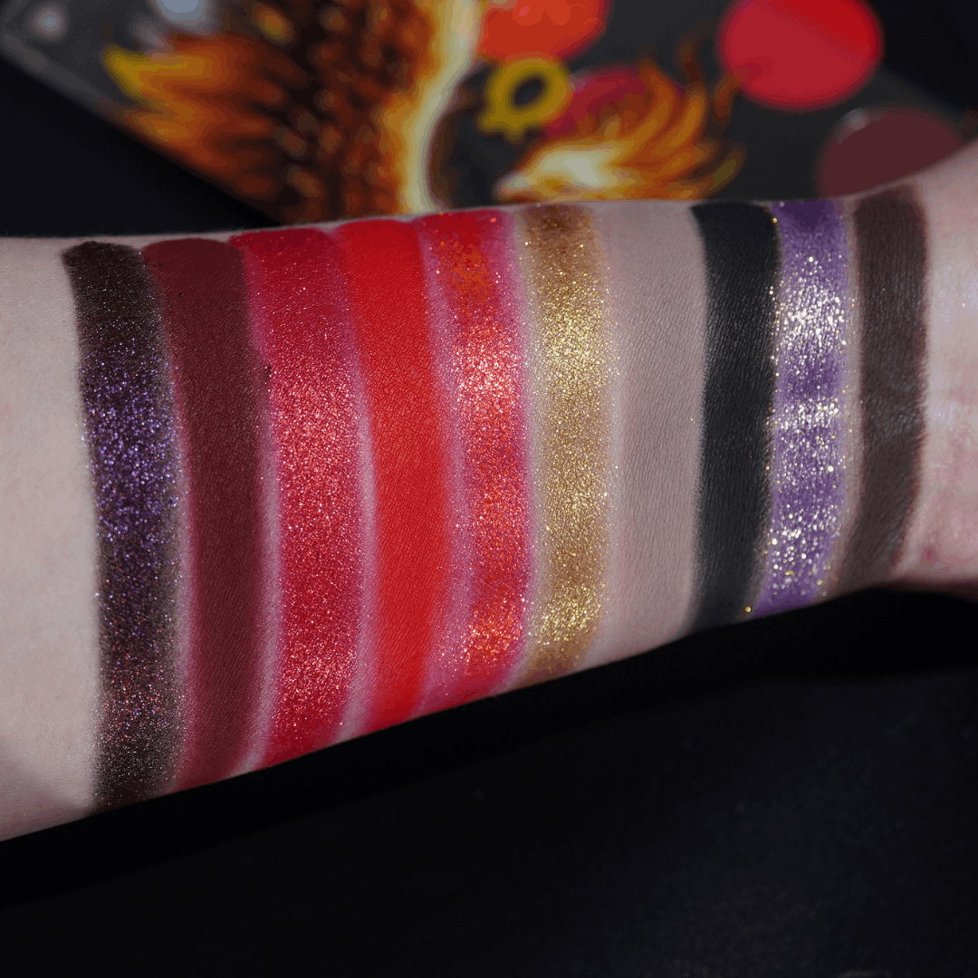 All ten shades swatched as wide stripes on a fair forearm, from purple sparkle and reds to gold, taupe, black and brown, lid art behind.