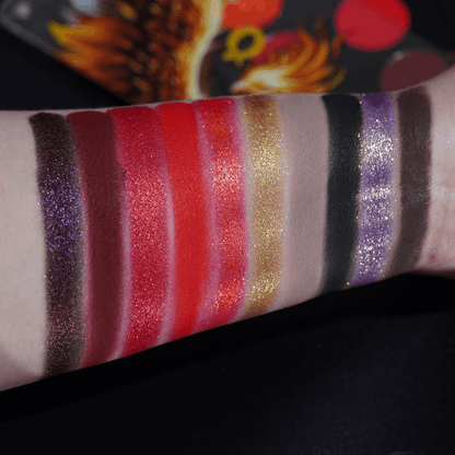 All ten shades swatched as wide stripes on a fair forearm, from purple sparkle and reds to gold, taupe, black and brown, lid art behind.