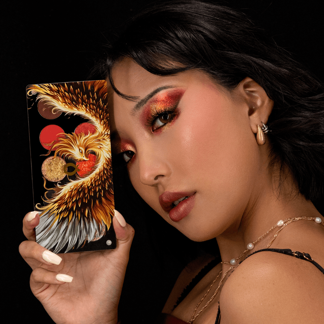 Model with a red and gold eye look holding the closed phoenix lid palette beside the face on a black background.