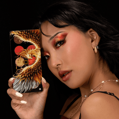 Model with a red and gold eye look holding the closed phoenix lid palette beside the face on a black background.
