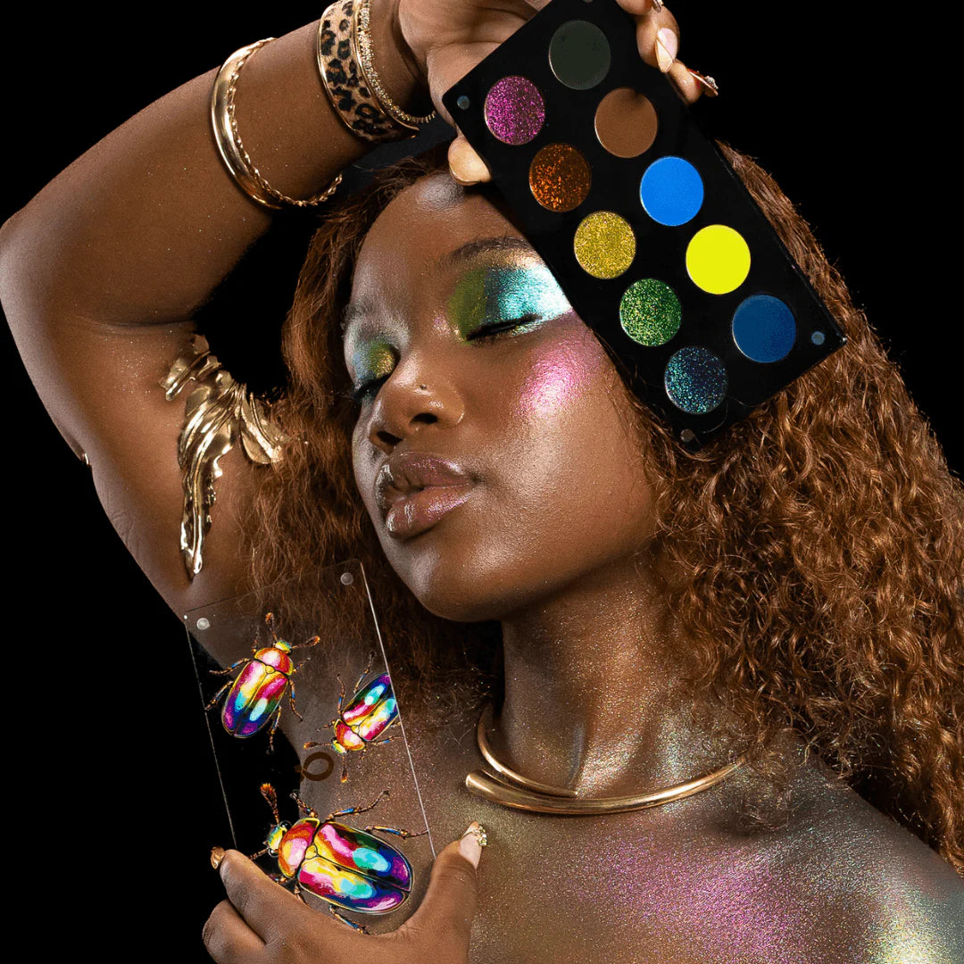 Question Beauty Garden of Iridescent Wings Chapter 4 - model holds eyeshadow palette, wearing multichrome green-blue eye look and gold jewellery, illustrated rainbow beetles in foreground, black background